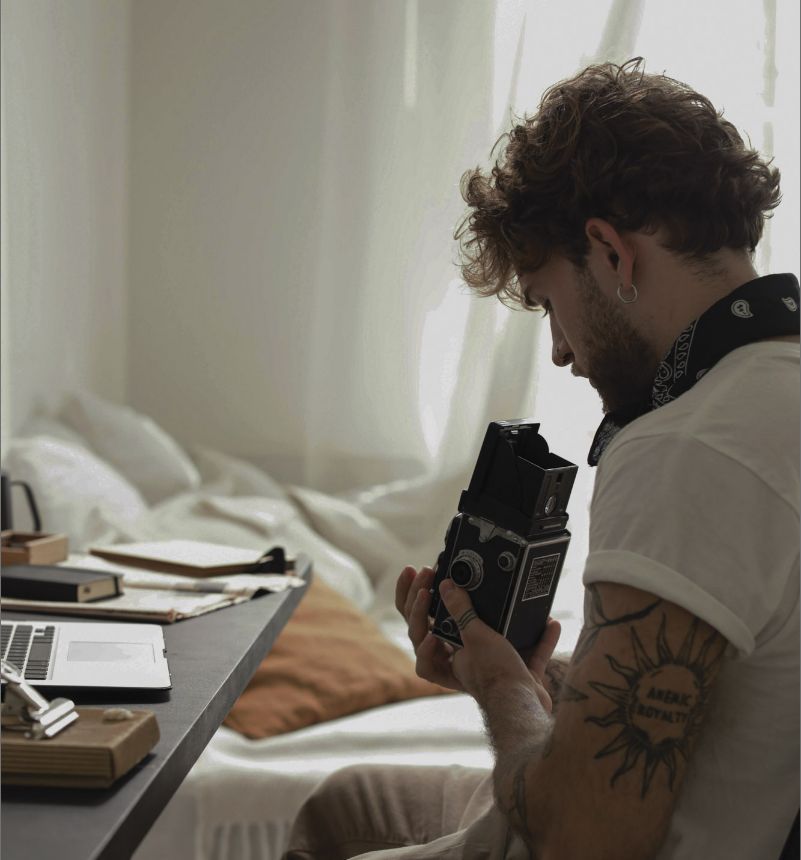 Man with arm tattoos sitting at a desk and holding a vintage film camera