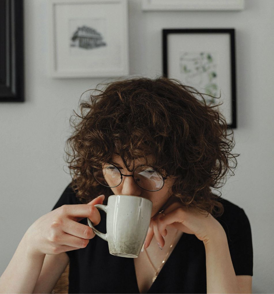 Close-up of a woman with curly hair and glasses drinking from a ceramic mug
