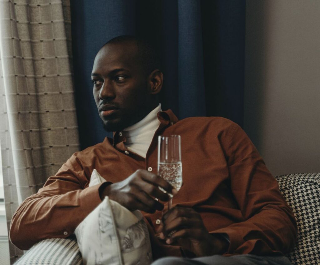 Man in a brown shirt and turtleneck sitting in a houndstooth armchair while holding a champagne flute